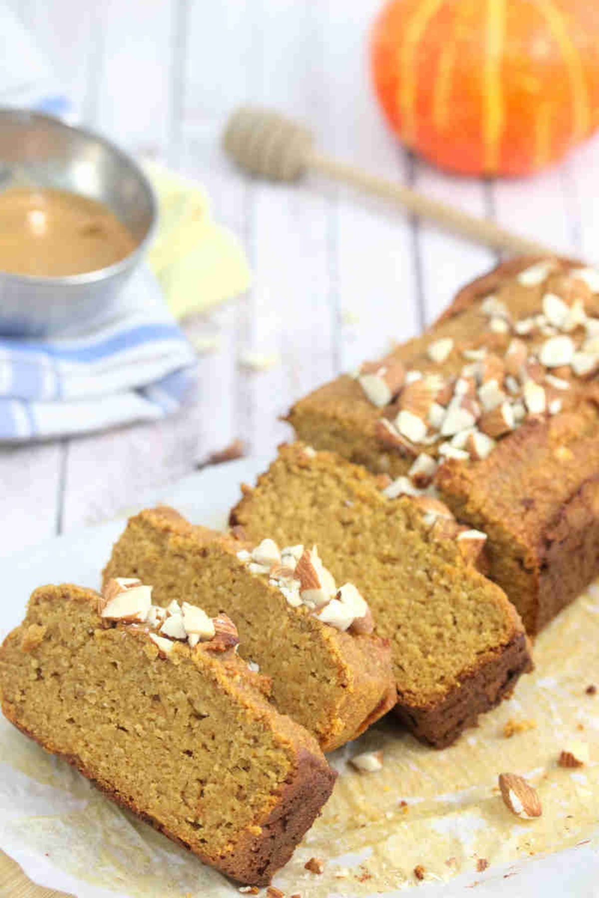 Pumpkin protein bread in slices on a wooden cutting board with almonds, linens, a pumpkin and honey stick around it.