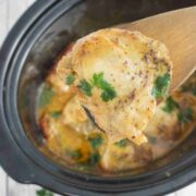 Close up of a wooden spoon holding a chicken thigh covered in honey mustard sauce with parsley over a black slow cooker.