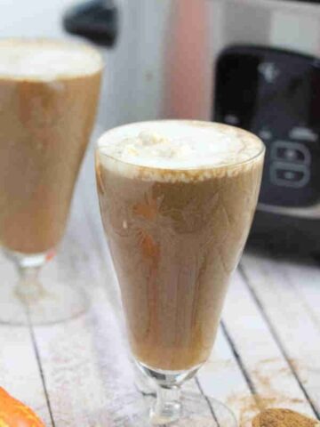 Two tall glasses with slow cooker pumpkin spice latte topped with coconut milk and pumpkins and a slow cooker around them.