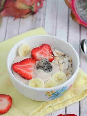 White bowl with a dragon fruit smoothie bowl in it topped with bananas, strawberries, and coconut on a yellow linen with berries, dragon fruit, and spoons around it.