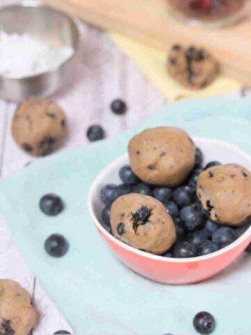 Pink bowl filled with blueberries and protein balls on a blue linen with more balls and dried blueberries around it.