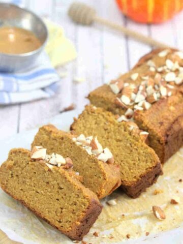 Pumpkin protein bread in slices on a wooden cutting board with almonds, linens, a pumpkin and honey stick around it.