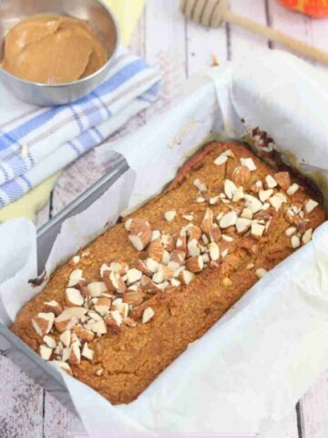 Pumpkin protein loaf baked in a pachment paper lined pan with peanut butter, a honey stick and linens around it.