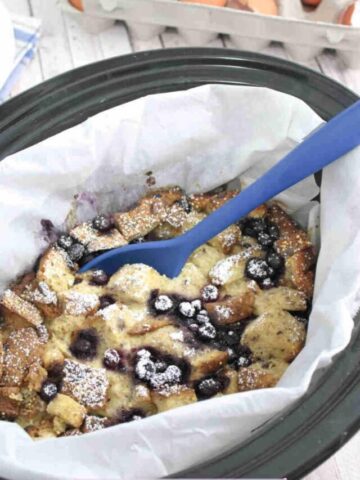Slow cooker lined with parchment paper and filled with high protein French toast casserole with blueberries with a blue spoon in the bowl.