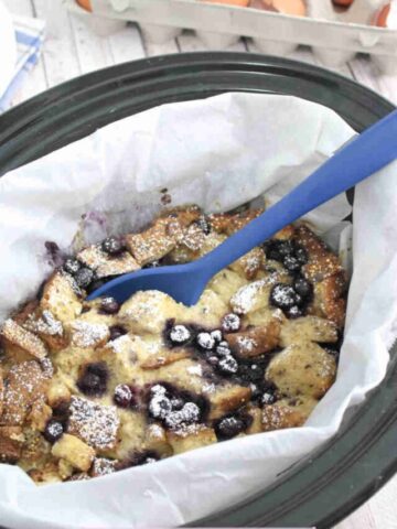 Slow cooker lined with parchment paper and filled with high protein French toast casserole with blueberries with a blue spoon in the bowl.