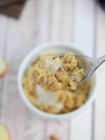Apple mug cake in a white ramekin with coconut milk on top and spoon pilling out of it.