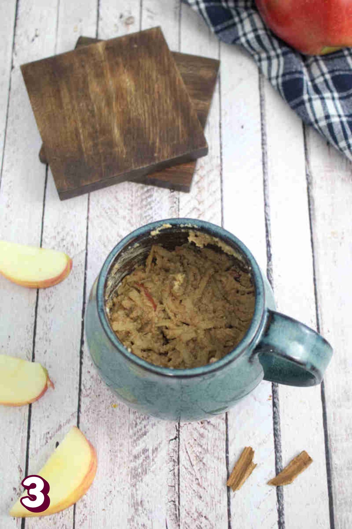 Blue mug with raw apple mug cake in it and sliced apples and cinnamon around it.