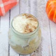 Mason jar with pumpkin spice overnight oats with cream cheese frosting on top and an orange linen and decorative pumpkins behind it.