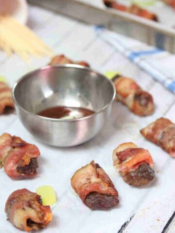 Close up of bacon wrapped steak bites on parchment paper with a steel bowl of bbq sauce.