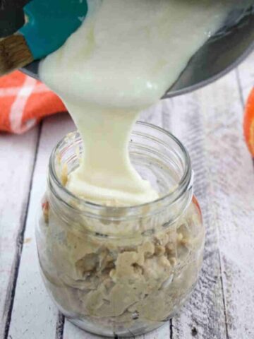 Mason jar with pumpkin spice overnight oats and cream cheese frosting being poured on top with an orange linen and decorative pumpkins behind it.
