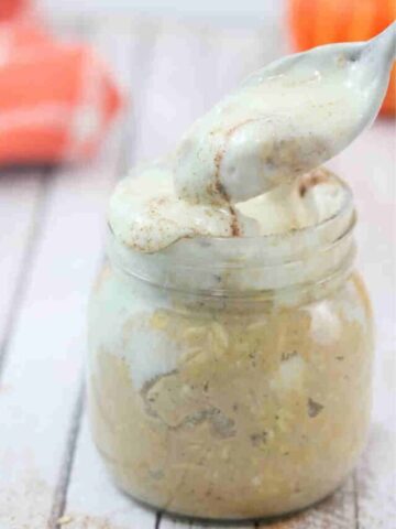 Spoon being pulled out of Mason jar with pumpkin spice overnight oats with cream cheese frosting on top and an orange linen and decorative pumpkins behind it.