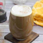 Dairy-free maple latte in a glass with milk frothed on top and the glass on a coaster with a yellow linen and french press behind it.