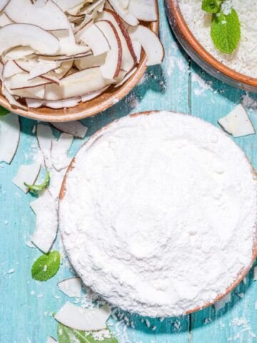 A bowl of coconut flour on a blue backdrop and more coconut.
