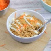 Carrot mug cake in a white ramekin with a spoon in the ramekin and cream cheese frosting on top. Ramekin on a cutting board with carrot behind it.