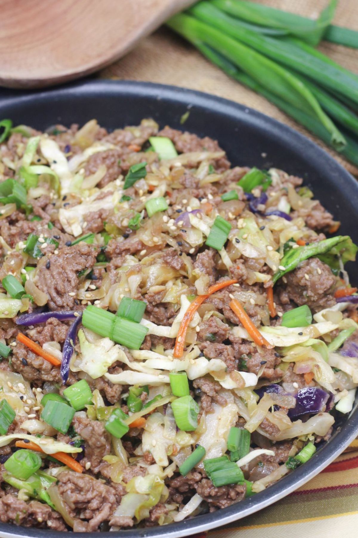 Inside out egg roll in a skillet with green onions on top and a wooden spoon and white plates behind it.