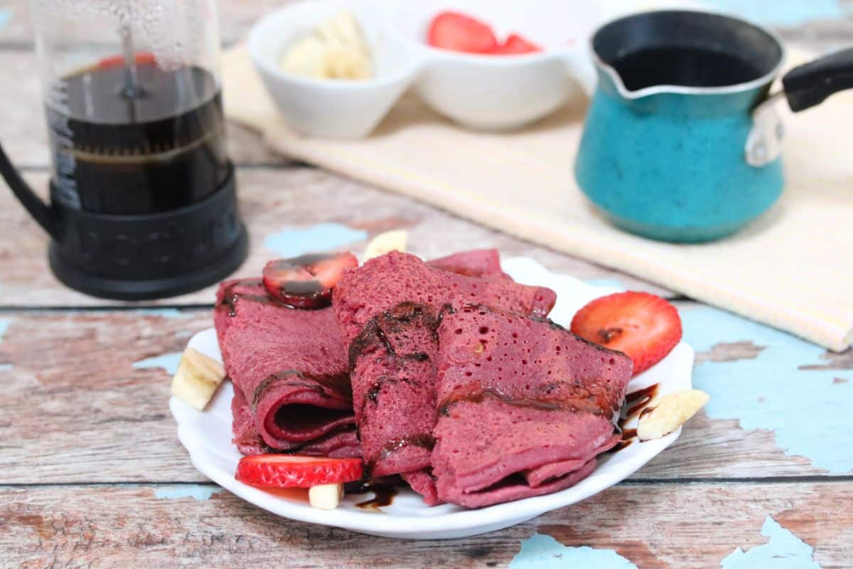 three pink vegan crepes on a white plate with chocolate, strawberries and banana on them and coffee in a french press behind them.
