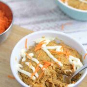 Carrot mug cake in a white ramekin with a spoon in the ramekin and cream cheese frosting on top. Ramekin on a cutting board with carrot behind it.