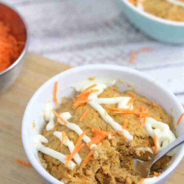Carrot mug cake in a white ramekin with a spoon in the ramekin and cream cheese frosting on top. Ramekin on a cutting board with carrot behind it.