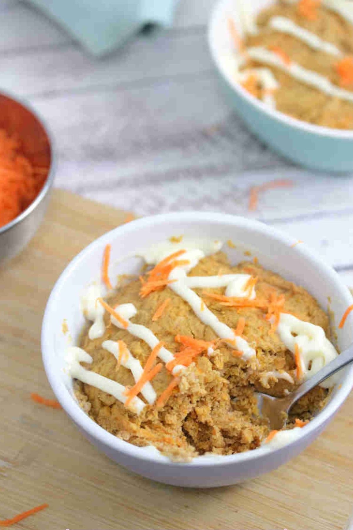 Carrot mug cake in a white ramekin with a spoon in the ramekin and cream cheese frosting on top. Ramekin on a cutting board with carrot behind it.