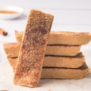 Snickerdoodle protein bars stacked with one bar in front and cinnamon sticks and milk behind it.
