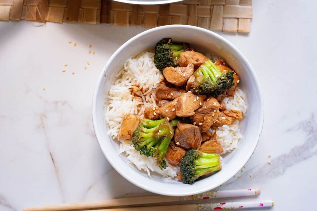 A bowl of white rice topped with cooked chicken pieces and broccoli florets. The dish is garnished with sesame seeds and placed on a light-colored marble surface. Wooden chopsticks with decorative handles are resting beside the bowl.