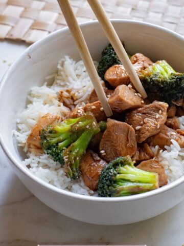 White bowl with teriyaki chicken pieces and broccoli with chopsticks beside it and chopsticks in the bowl.