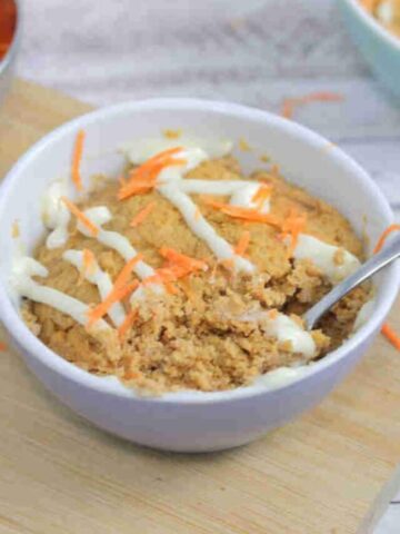 Carrot mug cake in a white ramekin with a spoon in the ramekin and cream cheese frosting on top. Ramekin on a cutting board with carrot behind it.