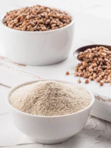 White bowl with buckwheat flour in it with buckwheat groats in a wooden spoon and another bol behind it.
