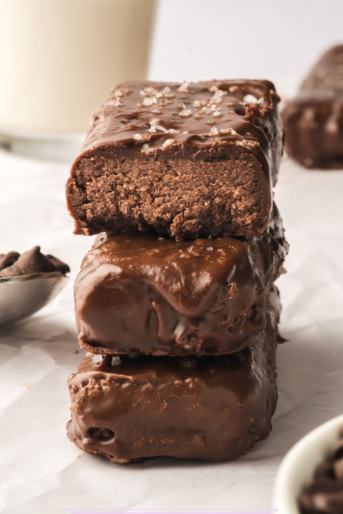 Three chocolate coated protein bars stacked on a white plate top bar with a bite out of it.