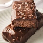 Two chocolate coated protein bars stacked on a white plate top bar with a bite out of it.