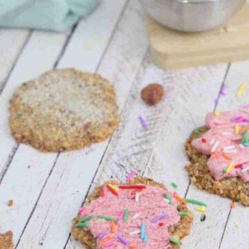 Sugar cookies with pink icing and sprinkles, a blue linen and bowl of sugar behind them.