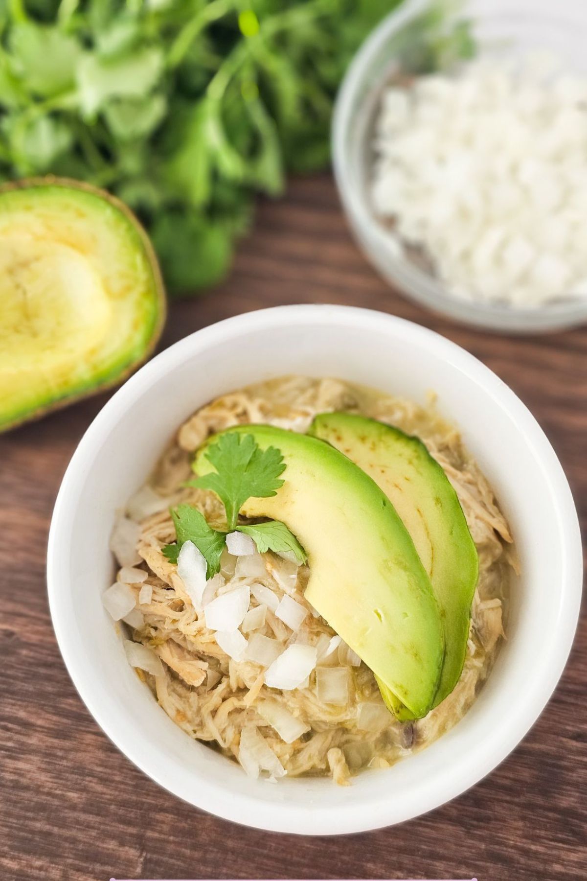 White ramekin topped with shredded chicken, onion and avocados on a cutting board.