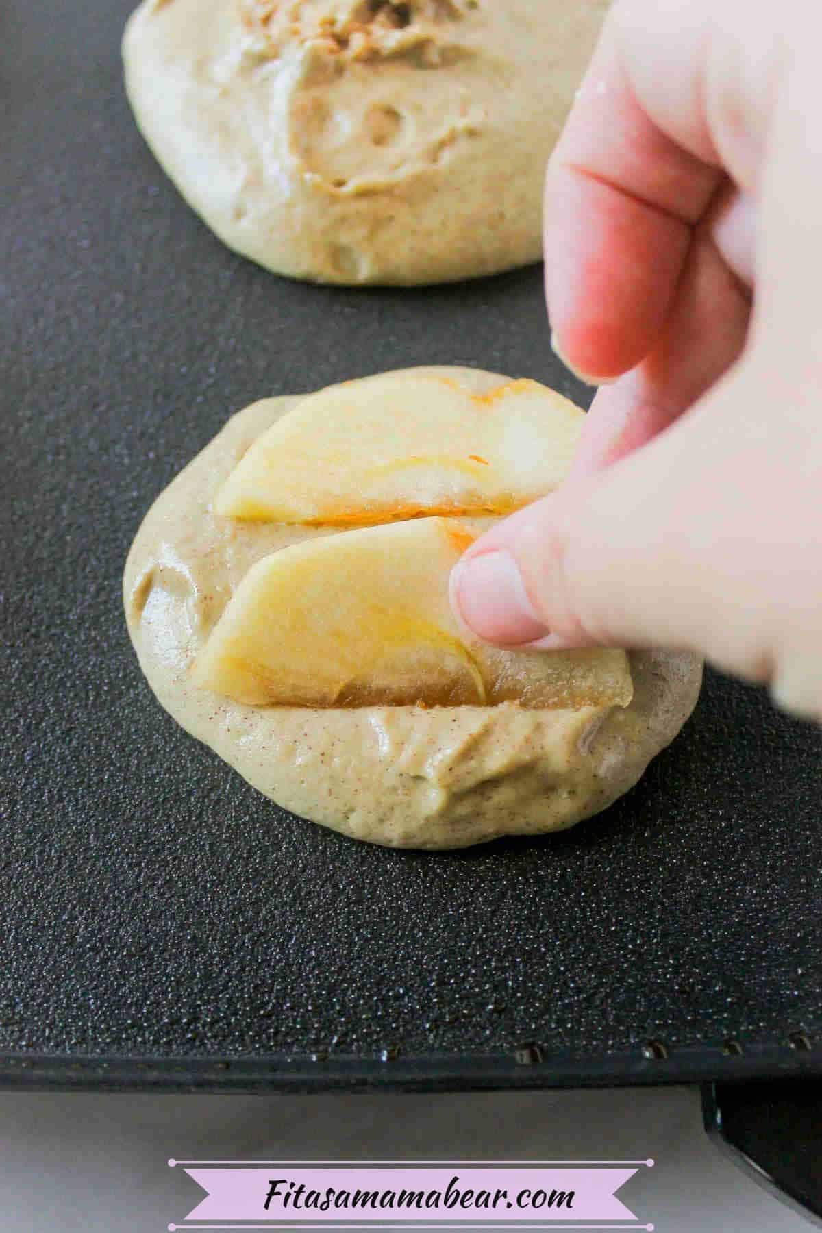 Apple sliced being put on raw pancakes on a pan.