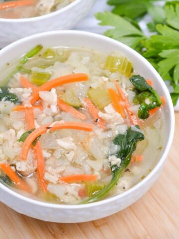 Chicken and cauliflower rice soup with carrots in a white bowl on a wooden cutting board.