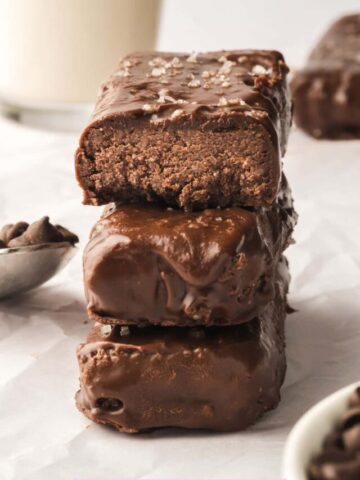 Three chocolate coated protein bars stacked on a white plate top bar with a bite out of it.