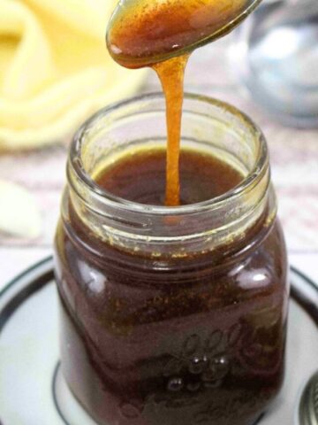 Gluten-free teriyaki sauce in a small jar on a white plate with garlic and a yellow linen around it