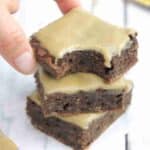 Three coffee brownies stacked on top of each other with the top one missing a bite and two fingers putting the brownie on.