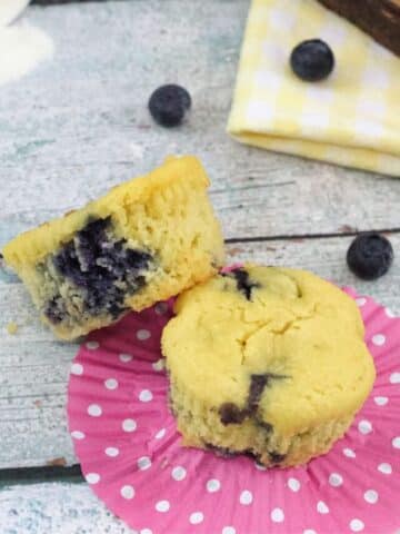 Two blueberry muffins on pink muffin liner with blueberries around it.