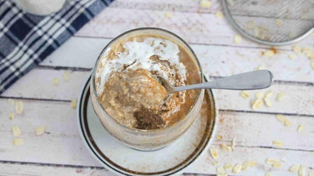 A glass jar of oatmeal topped with white yogurt or cream, sprinkled with cinnamon. A metal spoon is scooping out a portion from the jar. The jar is placed on a white wooden surface with scattered oats and a checkered cloth nearby.