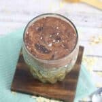 Clear glass with overnight protein oats in it topped with chocolate and sea salt on a wooden coaster on a blue linen.