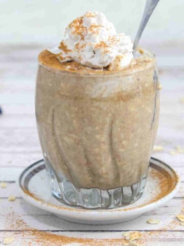 Blended overnight oats in a glass with whipped cream on top and a spoon in it and dusted with cinnamon.