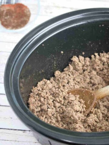 Cooked taco beef in a slow cooker with a wooden spoon in it.