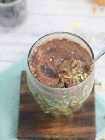 Clear glass with overnight protein oats in it topped with chocolate and sea salt on a wooden coaster on a blue linen with a spoon in the glass.