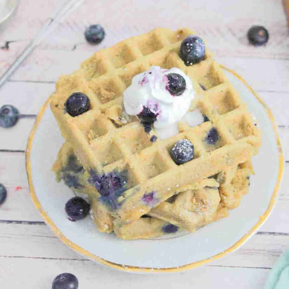 Three blueberry protein waffles stacked on a white plate topped with whip cream, blueberries and icing sugar.