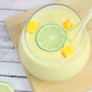 Mango protein smoothie in a glass on a cutting board topped with lime and mango and straws in the glass.
