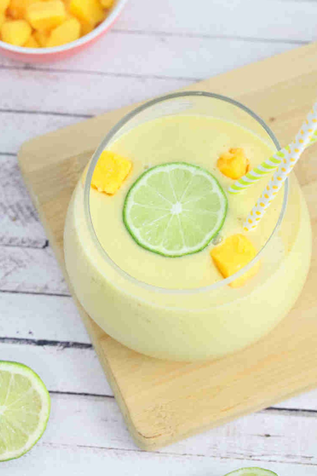 Mango protein smoothie in a glass on a cutting board topped with lime and mango and straws in the glass.