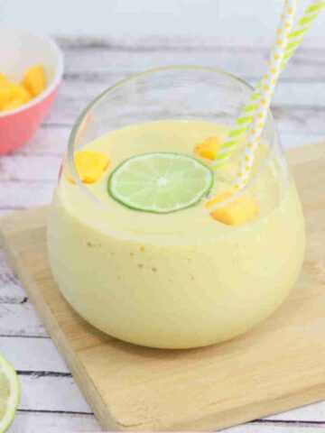 Mango protein smoothie in a glass on a cutting board topped with lime and mango and straws in the glass.
