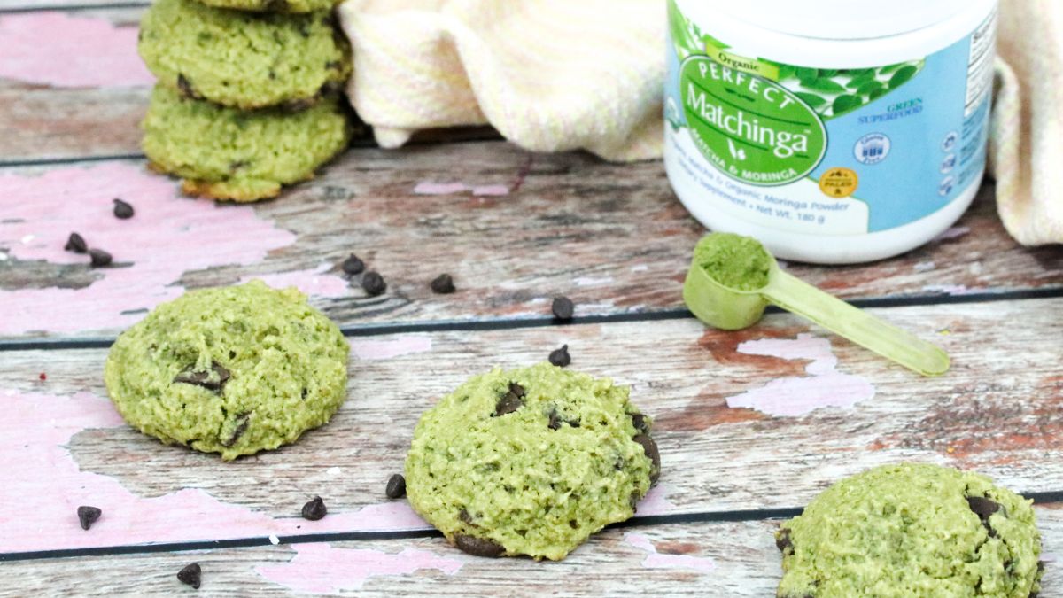 Three energy boosting cookies in a line with more stacked behind it and a tub of green matchinga powder with a scoop behind it.