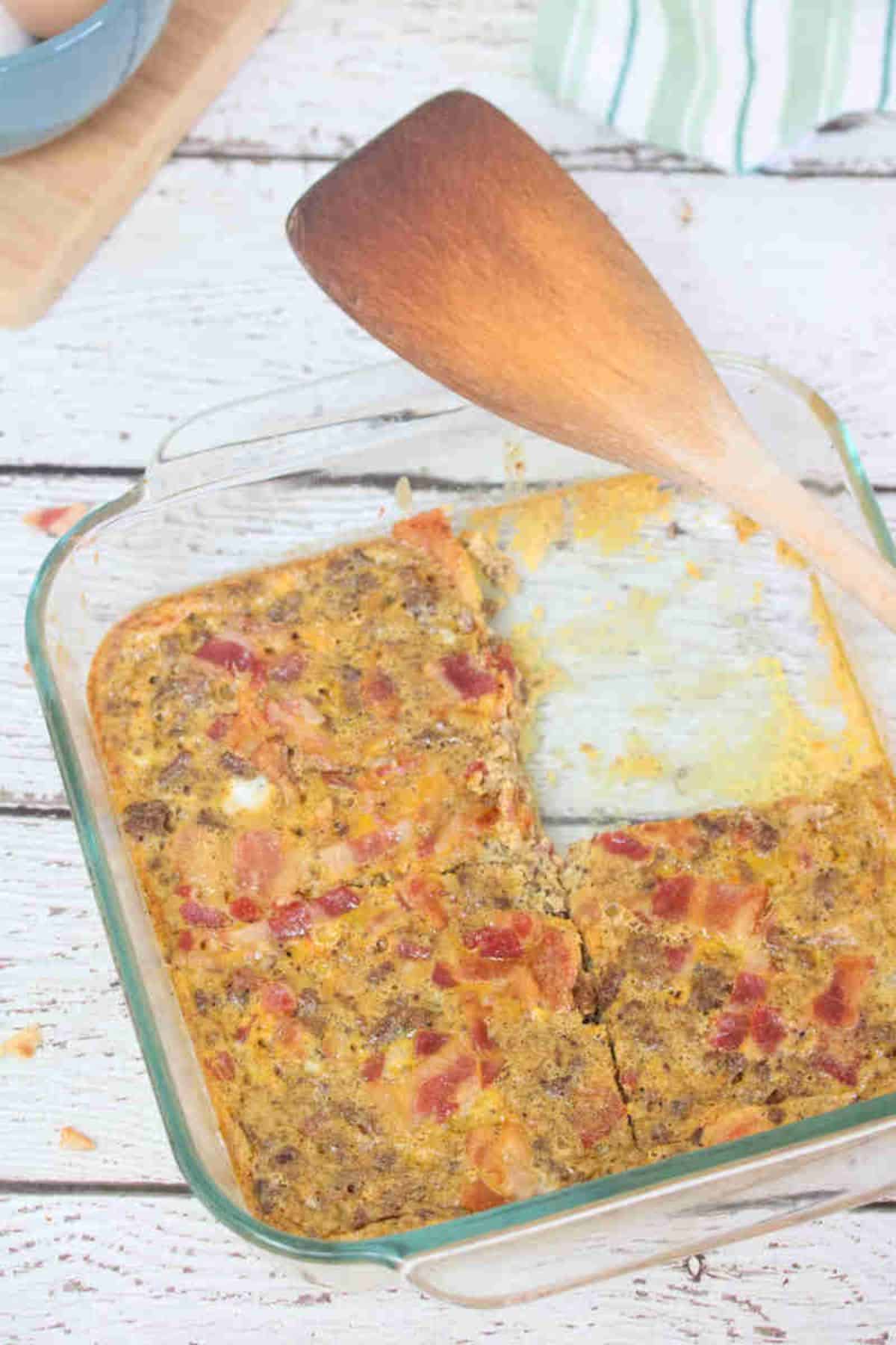 Carnivore casserole topped with bacon in a glass serving dish with a slice missing out of it and a wooden spatula on the side.