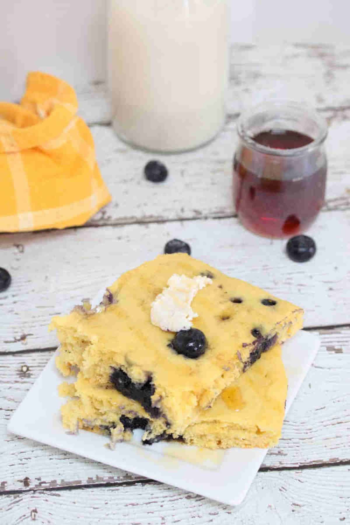 Protein pancakes with blueberries stacked on a white plate with maple syrup and milk behind them.